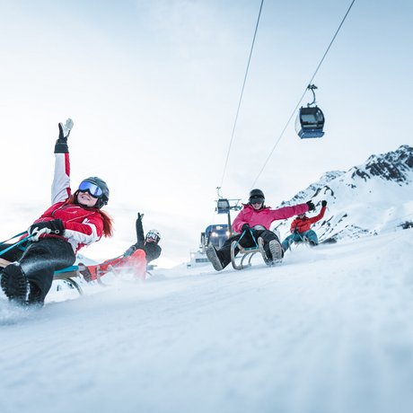 Ihre Unterkunft in St. Christoph am Arlberg: Bilder Vier Personen rodeln fröhlich einen verschneiten Berg hinunter mit Gondeln im Hintergrund