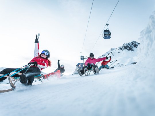 Hotel Maiensee: Wintersport am Arlberg Vier Personen rodeln fröhlich einen verschneiten Berg hinunter mit Gondeln im Hintergrund