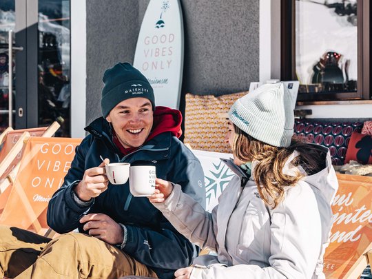 Das Maiensee: Ihr Hotel in St. Christoph Zwei Personen trinken heiße Getränke draußen auf der Terrasse im Schnee
