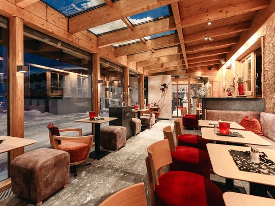 Die Lounge-Bar in Ihrem Skihotel am Arlberg Gemütliches Café mit Holzbalken und großen Fenstern, roter Dekoration und Sitzgelegenheiten.