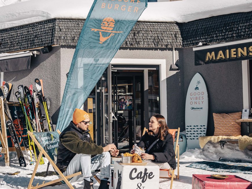 Burger im Skihotel in St. Anton am Arlberg Zwei Personen sitzen draußen im Schnee vor einem Café und Bar, umgeben von Skiern.